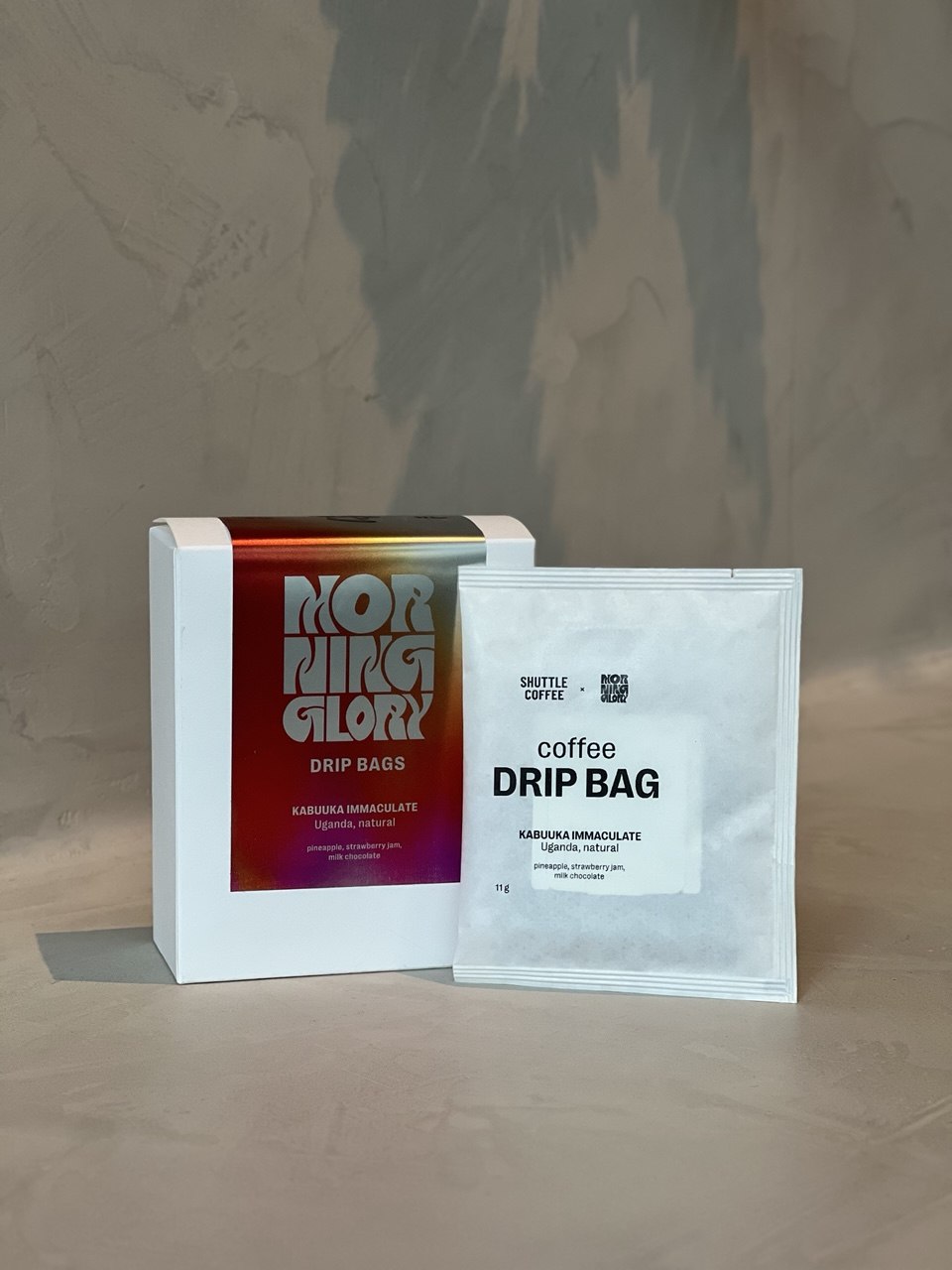 A white box labeled MORNING GLORY DRIP BAGS next to a single white drip bag packet for coffee, set on a beige surface against a textured beige wall.