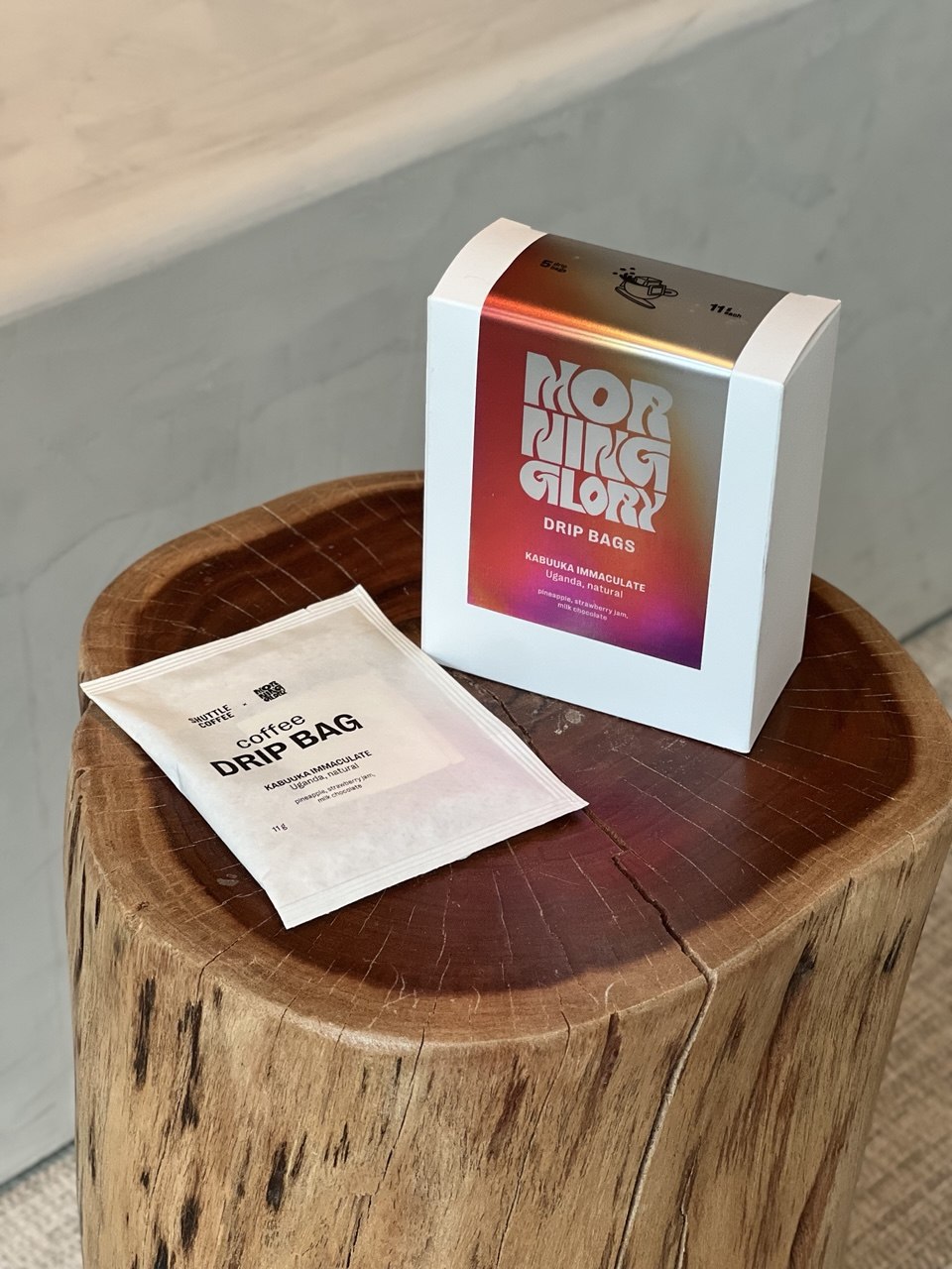 A white box labeled 'Morning Glory Drip Bags' and a small white drip bag sit on a round wooden stump in an indoor setting.